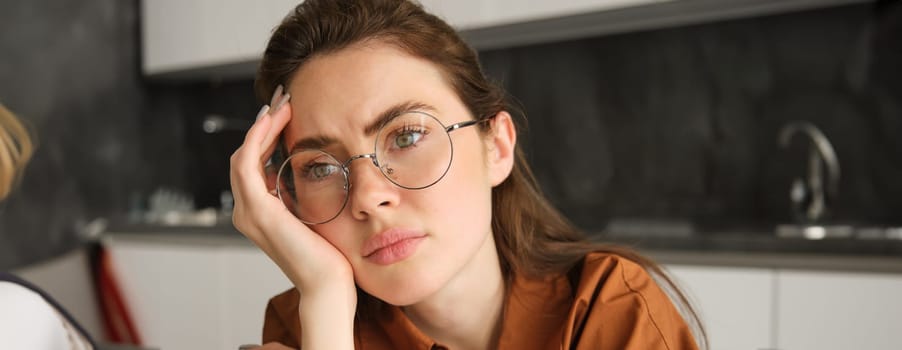 Portrait of sad and tired young woman in glasses, sitting in kitchen at home, looking upset and exhausted, working from home.