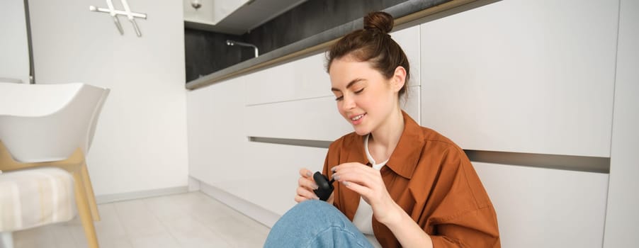 Portrait of young smiling woman, girl puts on her black wireless headphones, listens to music in earphones, sits on kitchen floor at home.