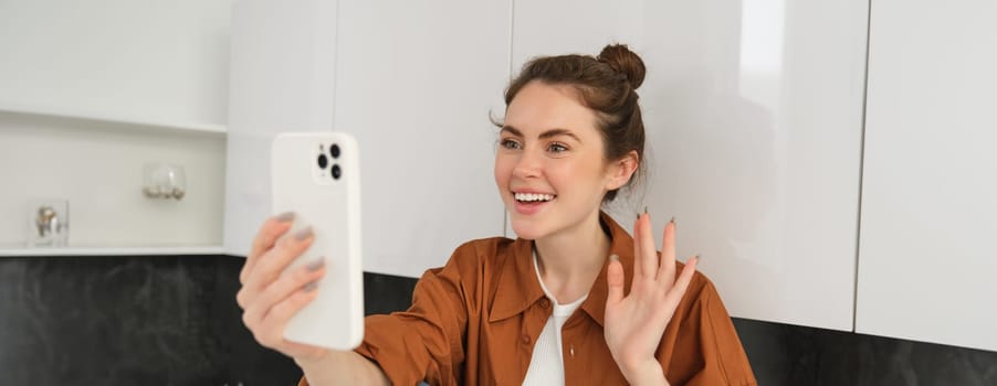 Portrait of young cheerful woman laughing and smiling during phone call, video chats with friend, sits on kitchen counter and talks to someone using smartphone app.