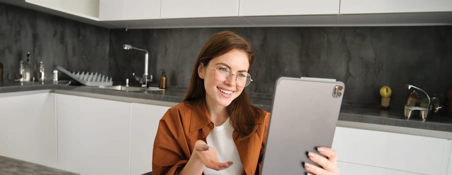 Portrait of young woman joins online course on digital tablet, having conversation, chatting on remote, sitting in kitchen.