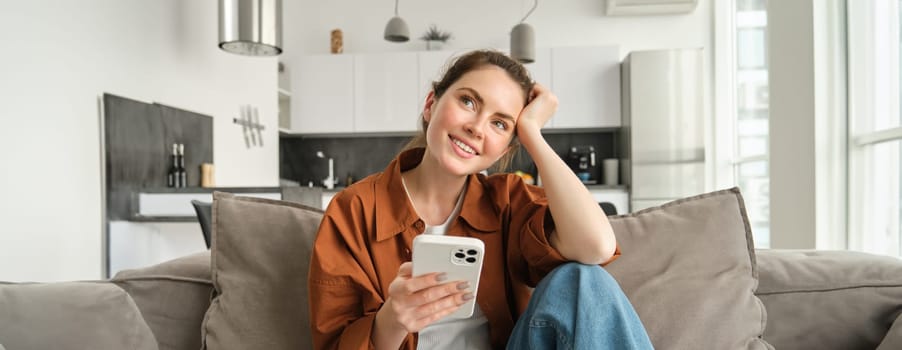 Smiling brunette woman sitting on sofa with smartphone, looking thoughtful, thinking while using mobile phone app, ordering on application, doing online shopping and resting at home.