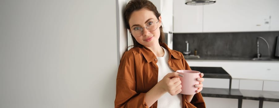 Portrait of modern woman at home, relaxing in her living room, leaning on wall, holding cup of tea and smiling, wearing glasses, drinking coffee and resting.