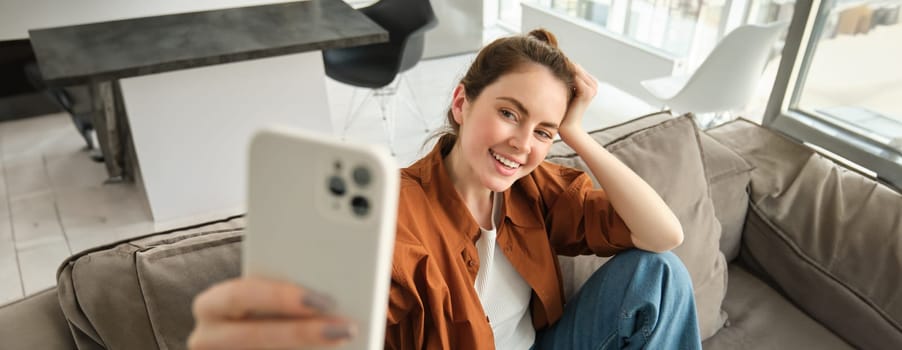 Portrait of beautiful happy girl takes selfies on sofa. Woman posing for photos on her couch, taking pictures for social media, extends hand with smartphone.