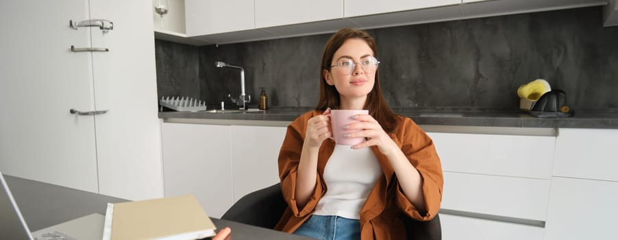 Portrait of young professional, girl freelancer sitting in kitchen and enjoying cup of coffee, sitting with documents, taking break from studying and smiling, looking outside window.