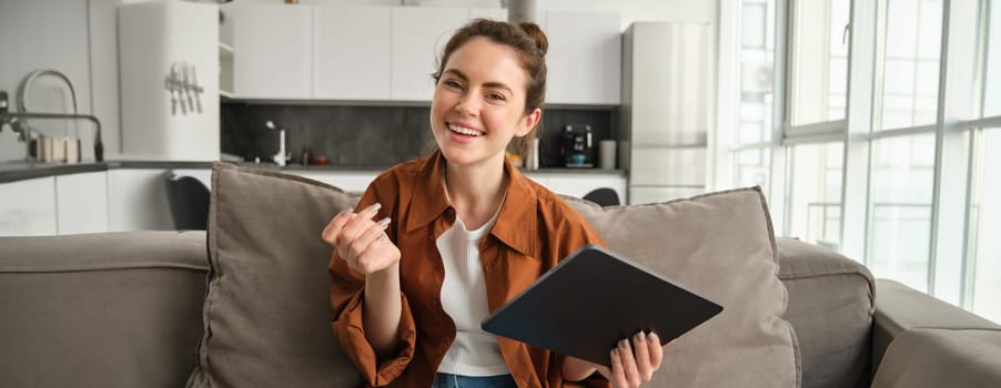 Portrait of smiling, carefree brunette woman, sitting on couch in living room, online shopping or reading on digital tablet, looking happy at camera.