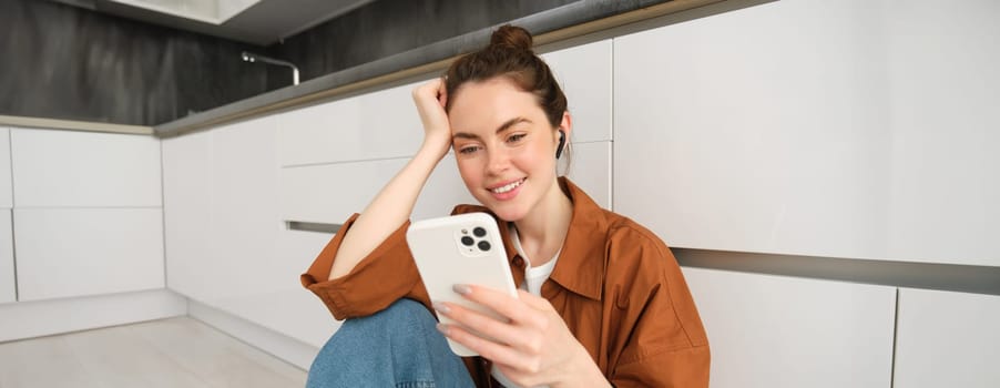 Portrait of happy young beautiful woman, listening music in wireless earphones, using smartphone, watching videos on mobile phone with headphones on.