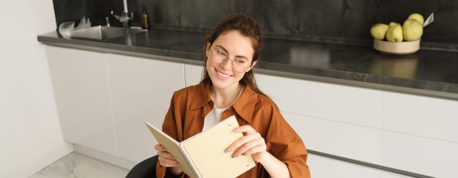 Close up portrait of young woman, student in kitchen, holding notebook, revising for exam at home, studying, reading her planner.