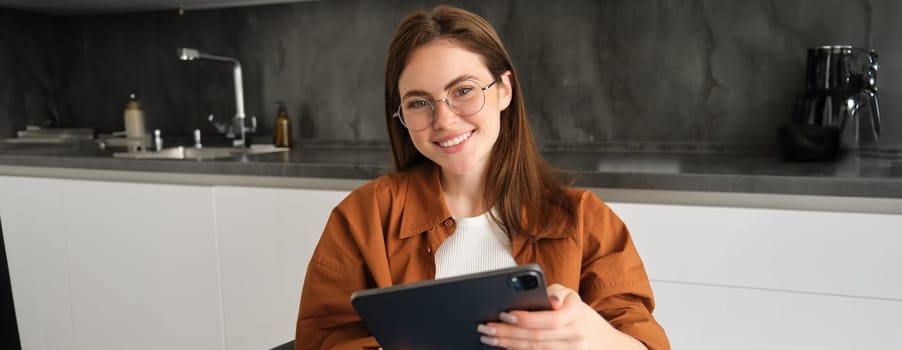 Portrait of beautiful brunette woman in glasses, sitting at home, holding digital tablet, smiling at camera, working remote, studying, giving lessons on online platform.
