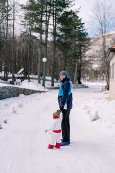 Smiling dad with a small child are standing on a snowy road near the house, holding hands. Side view. High quality photo