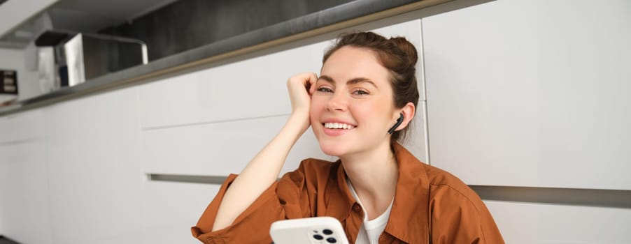 Carefree smiling woman with smartphone, messaging, using mobile phone, sitting on kitchen floor, listening music in wireless headphones.