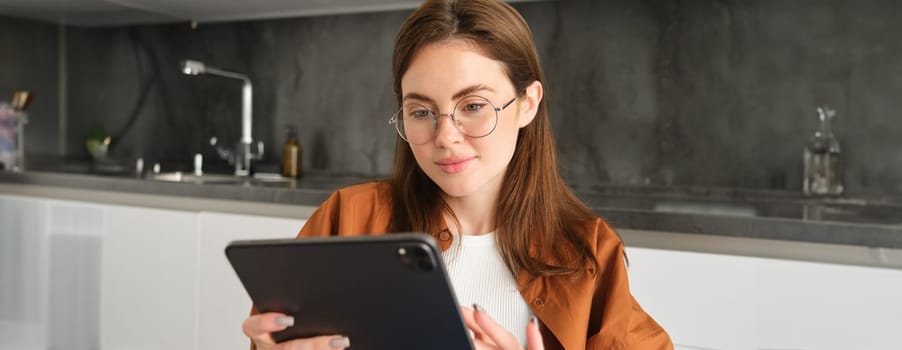 Portrait of busy young woman, self-employed entrepreneur sitting at home with digital tablet, reading documents. Student working on project remotely, holding gadget, studying.