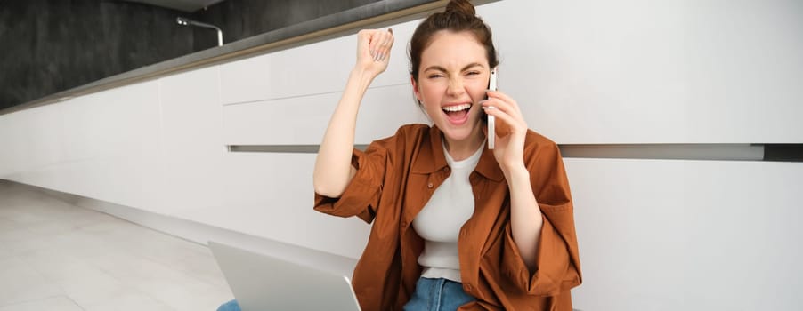 Enthusiastic young woman celebrating, talking on mobile phone, sits on floor at home with laptop and feeling excitement, winning or triumphing.