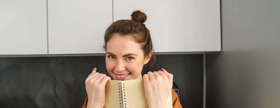 Portrait of young brunette woman in the kitchen, holding notebook, reading notes on planner, laughing and smiling.
