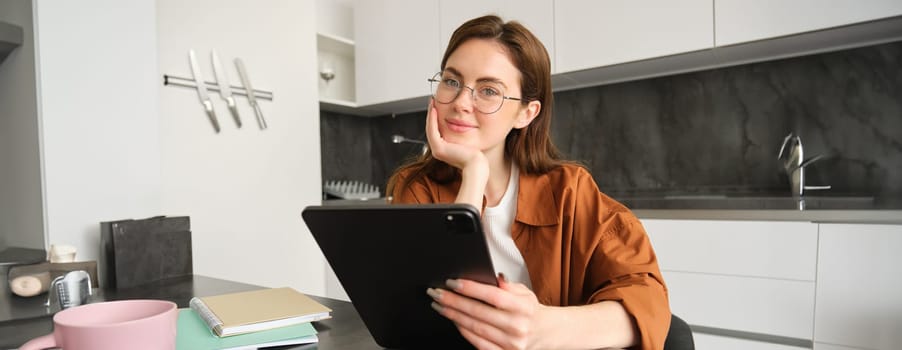 Portrait of cute young woman, freelance teacher, sitting in kitchen wearing glasses, holding digital tablet, teaching people online, using gadget to connect to lesson, working remotely.