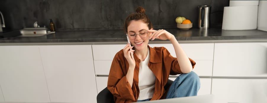 Image of beautiful modern woman working from home, studying in kitchen with laptop, talking on smartphone, calling someone on telephone.