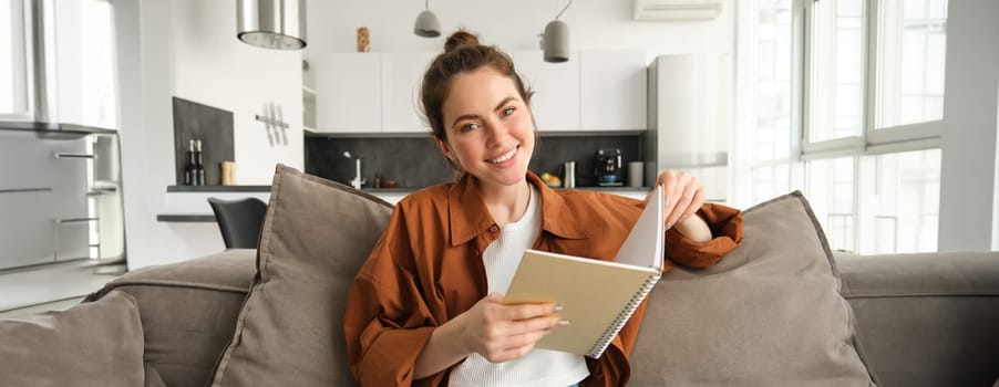 Young woman sits at home on sofa, reading her notes, holding notebook, studying for exam, student revising in her living room.