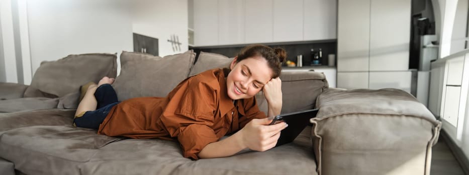 Portrait of beautiful young woman resting at home on couch, lying on sofa with digital tablet, reading or watching videos on gadget.