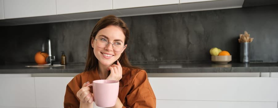 Portrait of young brunette woman in kitchen, resting indoors, smiling and drinking coffee, enjoys tasty hot tea.