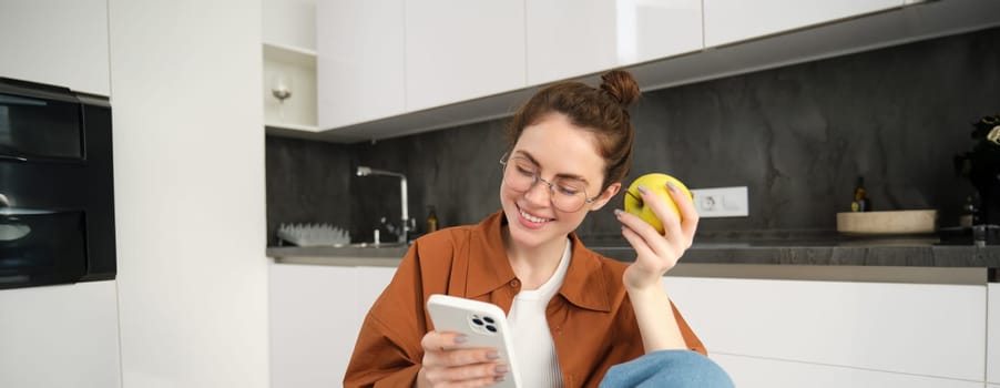 Young woman chilling at home, looking at smartphone, reading news on social media and eating green apple in the kitchen.