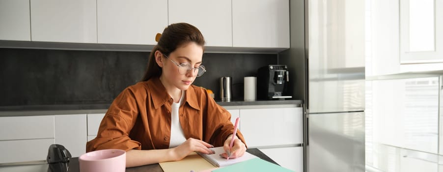 Portrait of woman looking concentrated, writing down notes, doing homework in kitchen, drinking coffee, studying at home.