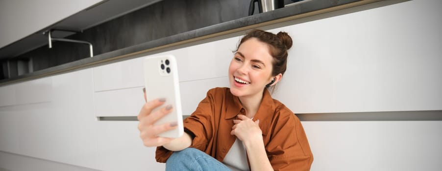 Portrait of beautiful smiling woman, talking to friend on smartphone video chat, connects to online conversation on mobile app, sitting on kitchen floor.