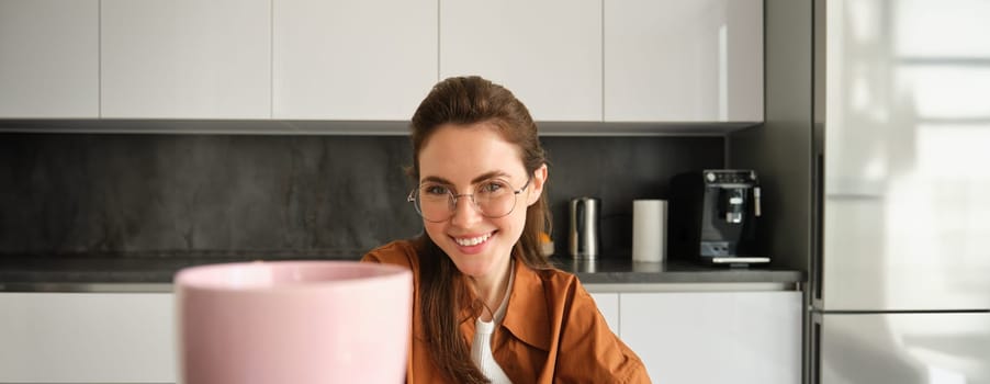 Beautiful young woman in kitchen, giving you cup of tea, taking break and drinking coffee at home, wearing glasses, smiling at camera.