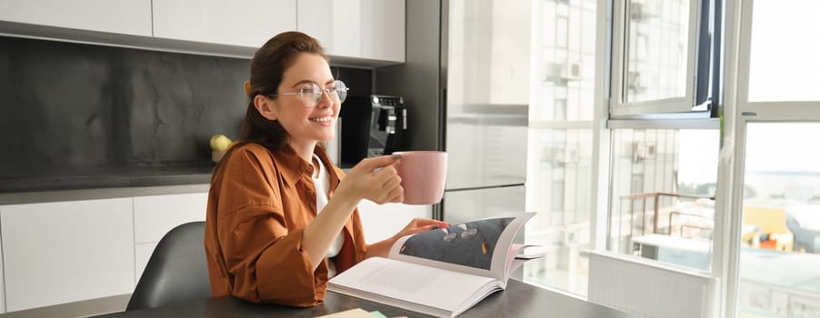 Relaxing day at home. Happy young woman in glasses, sitting with cup of tea or coffee, reading a book and smiling, studying, revising for lesson.