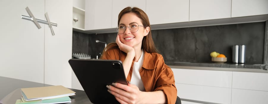 Portrait of cute woman, student in glasses, sitting in kitchen with digital tablet, working remotely, making home as workplace, teacher giving lessons online.