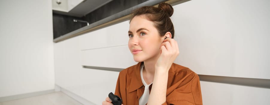 Portrait of beautiful woman enjoys listening to music or podcast in wireless black headphones, using earphones, smiling and looking happy, sitting at home on kitchen floor.