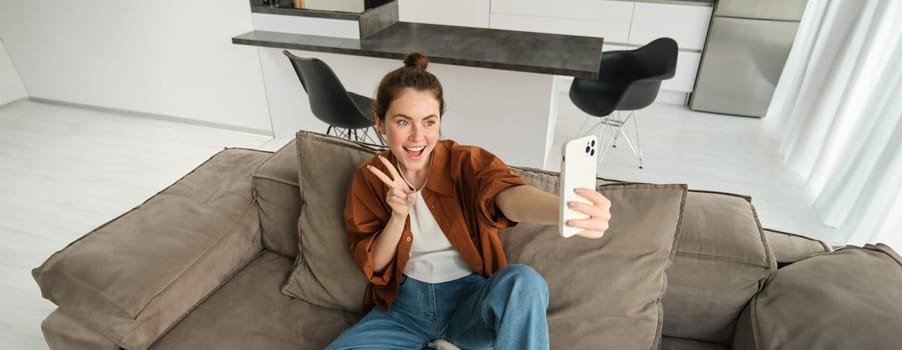 Portrait of cute smiling female model, takes selfie on smartphone app, posting photos on social media, using mobile phone camera, posing on sofa at home.