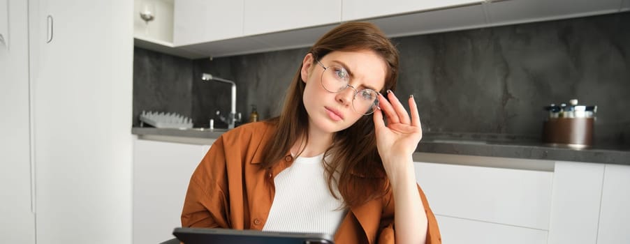 Portrait of serious young woman in glasses, holding tablet, sitting in kitchen, looking away in deep thoughts, thinking, has concerned face.