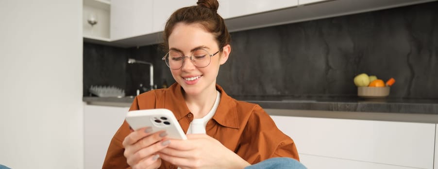 Portrait of young brunette woman in glasses, sitting at home with mobile phone, using her smartphone and smiling, reading message.