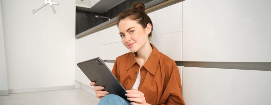 Portrait of young beautiful brunette woman, sitting on kitchen floor at home, holding digital tablet, smiling and looking at camera with happy face.