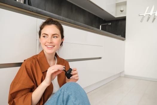 Image of modern brunette girl, sits on kitchen floor, listens to music in wireless headphones, holding earphones and case in hands, looking aside.