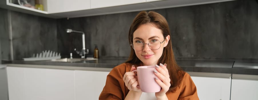 Young woman enjoying brewed coffee in kitchen, holding cup of her favourite drink at home and smiling.