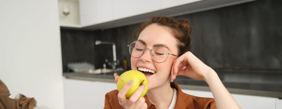 Portrait of smiling, modern woman in glasses, sitting at home and eating green apple, having a lunch break, biting fruit.