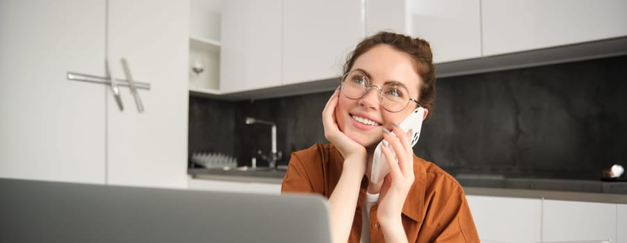 Portrait of young woman, business owner working from home, student making a phone call, sitting in kitchen with laptop, talking to someone.