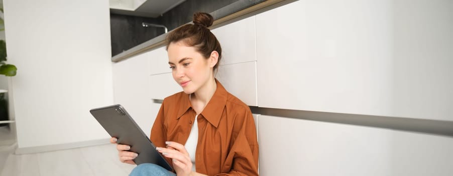 Portrait of pretty young woman at home, sitting on kitchen floor, looking at digital tablet, using gadget, reading on device.