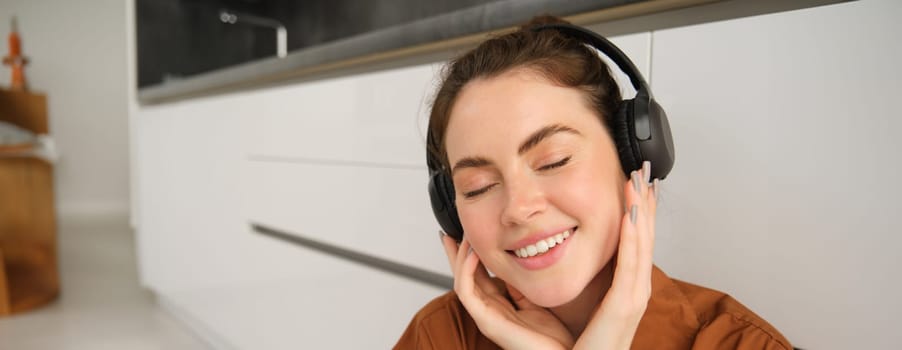 Close up portrait of beautiful brunette woman, listens to music in wireless headphones, has new earphones on her head, smiling and looking happy.