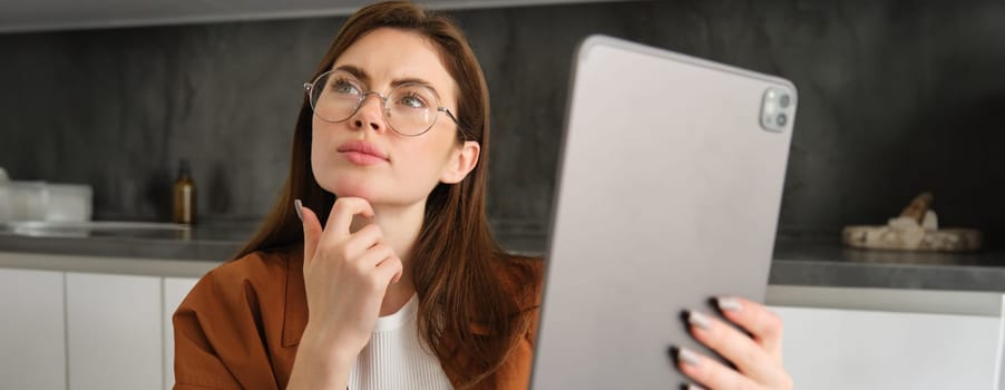 Close up portrait of serious woman working from home, holding digital tablet, looking aside with thinking face, pondering, studying online, works on remote.