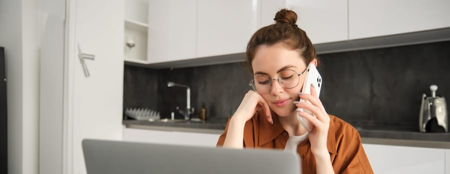 Close up portrait of young woman online shopping on laptop, working from home on computer, making a phone call and smiling, saleswoman talking to customer, works on remote.