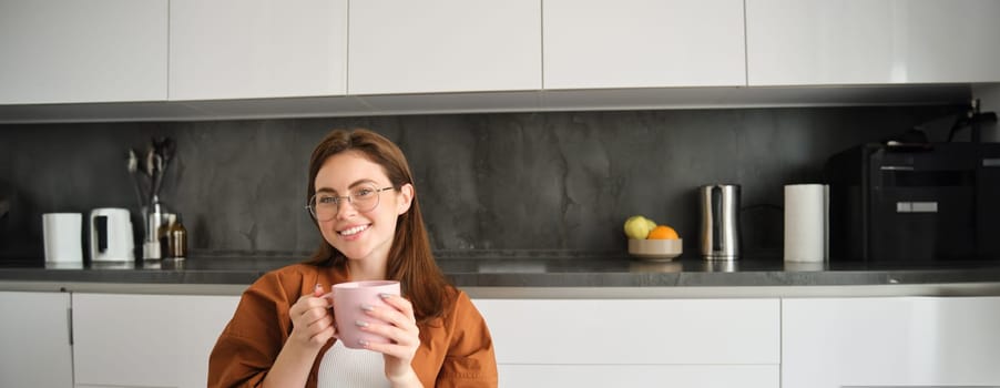 Portrait of happy young woman in glasses, sitting at home in kitchen, holding cup of coffee, drinking favourite tea, relaxing indoors on cold autumn day.