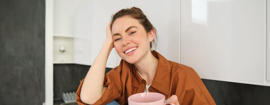 Beautiful woman offers you her cup of coffee, smiling and looking happy, sitting in kitchen, enjoying her morning drink.