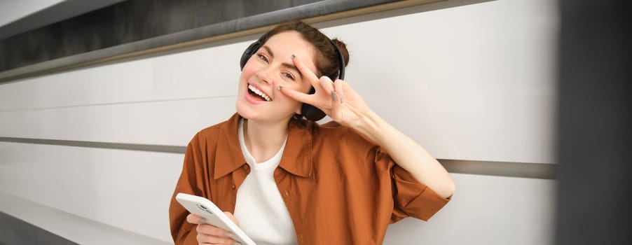 Positive young woman, sits at home with wireless headphones and smartphone, smiles and looks happy.