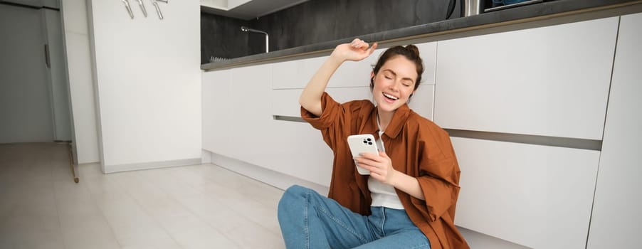 Portrait of woman enjoying watching videos on smartphone with wireless earphones, girl listening to music and using mobile phone, sitting on kitchen floor.