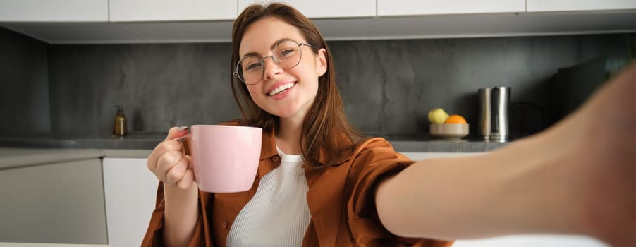 Portrait of happy young woman in glasses, takes selfie with smartphone, drinks coffee, posing with delicious mug of tea in kitchen.