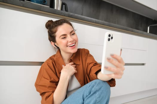Portrait of beautiful smiling woman, talking to friend on smartphone video chat, connects to online conversation on mobile app, sitting on kitchen floor.