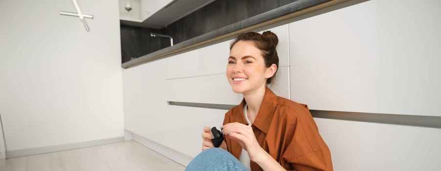 Portrait of young smiling woman listens to music in her black wireless earphones, using headphones to enjoy sound quality of song, sits on kitchen floor.