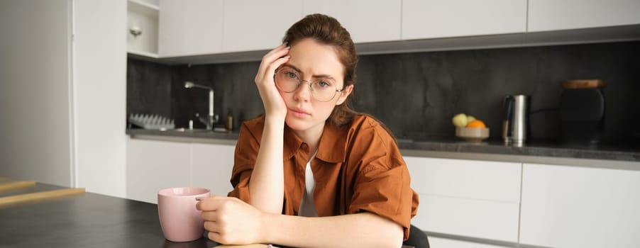 Portrait of young serious woman, working from home in glasses, sitting with folder and documents in kitchen, looking tired.