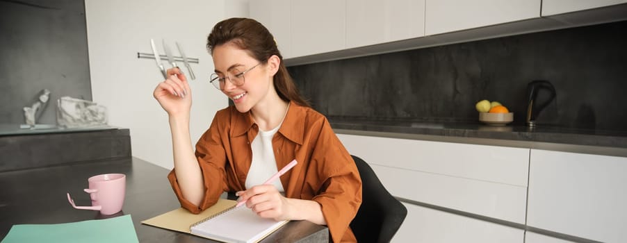 Portrait of young woman in glasses studying at home, writing down something in notebook, making notes, student doing homework in kitchen.